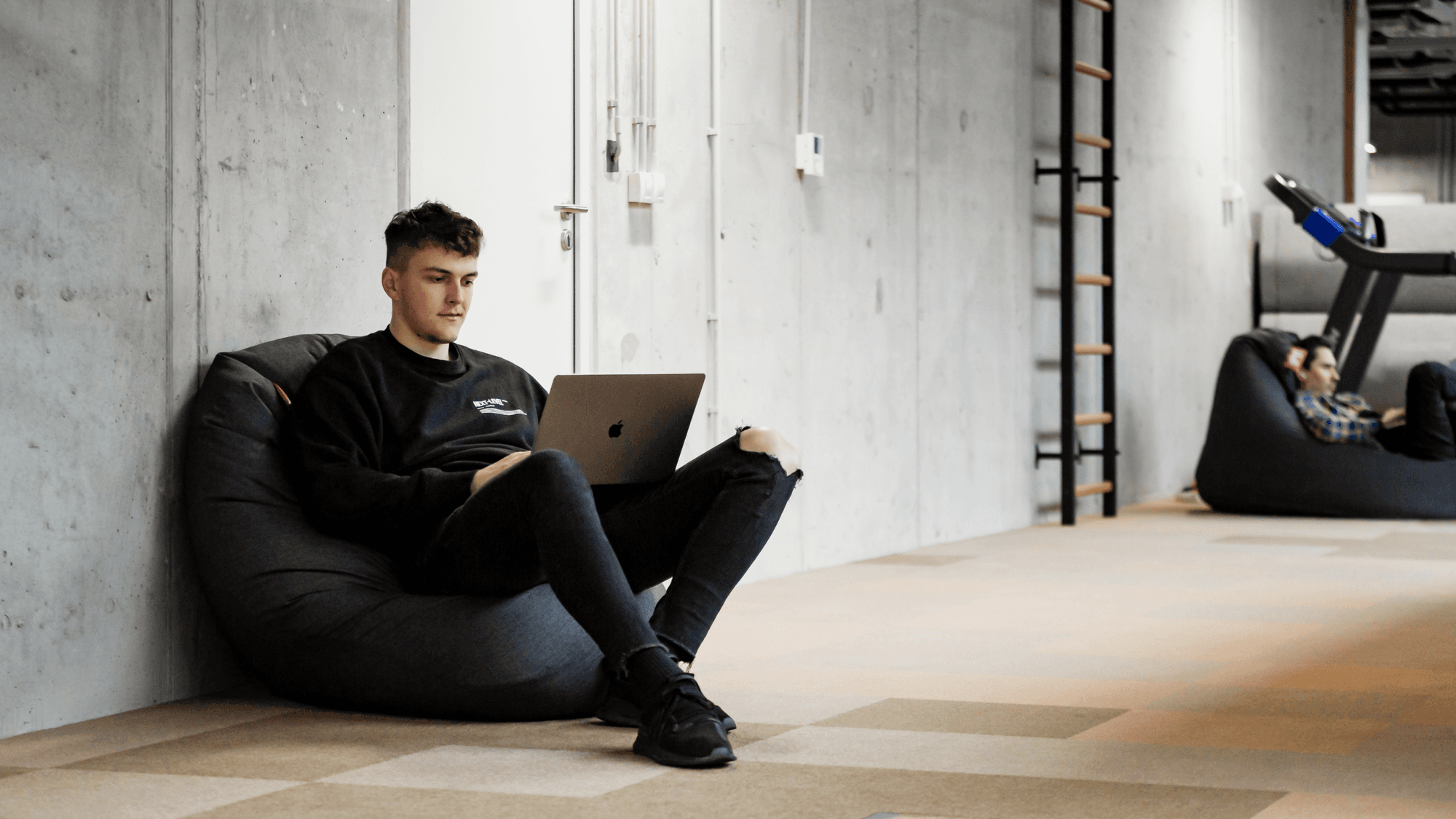 A young man sits on a bean bag, using a laptop in a modern, minimalistic workspace, with another person in the background.