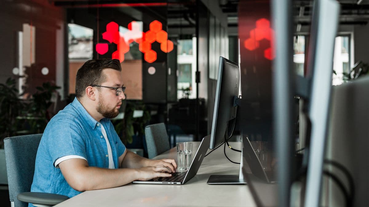 A man with glasses types on a laptop at a modern office desk, with vibrant red lights in the background.