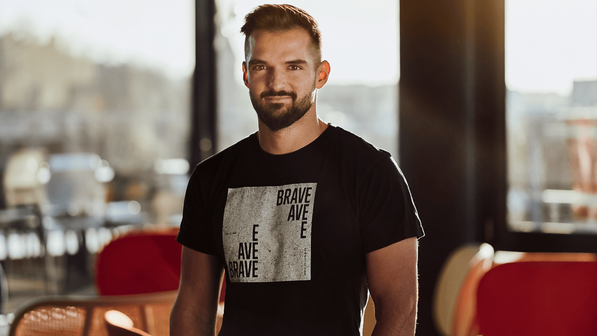 A man with a beard stands confidently in a sunny café, wearing a black t-shirt with "BRAVE AVE" printed on it.