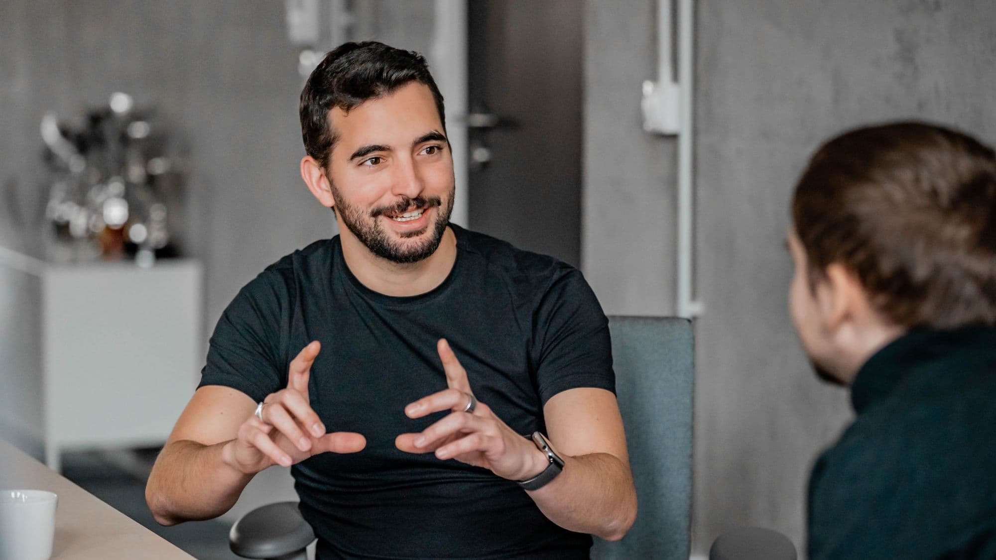 A man with a beard and short hair gestures emphatically while speaking to another person in a modern office setting.