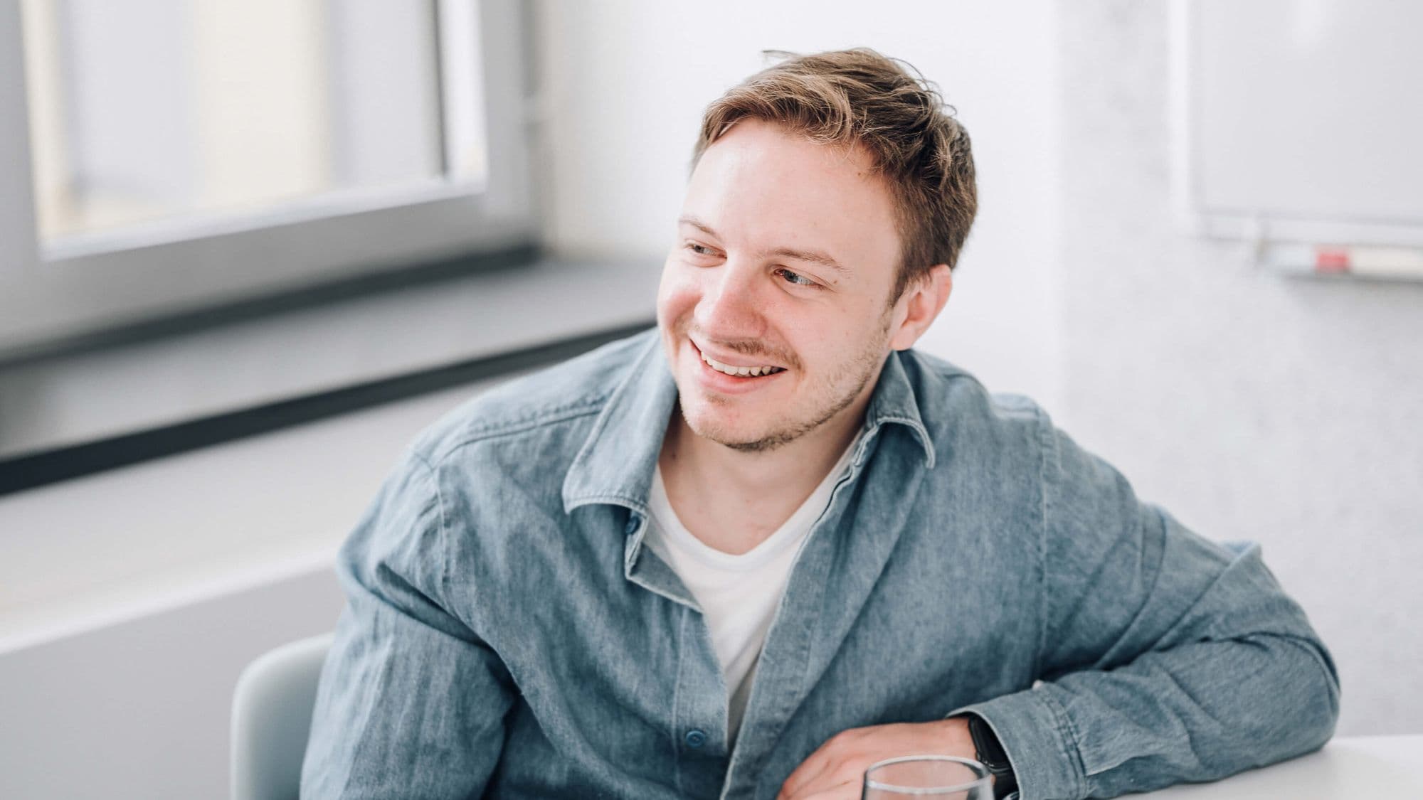 A smiling man with short hair, wearing a denim shirt, sits at a table with a glass in a bright room.