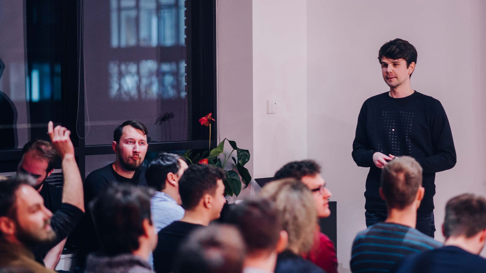 A speaker presents to an engaged audience in a bright, modern setting, with one attendee raising their hand.