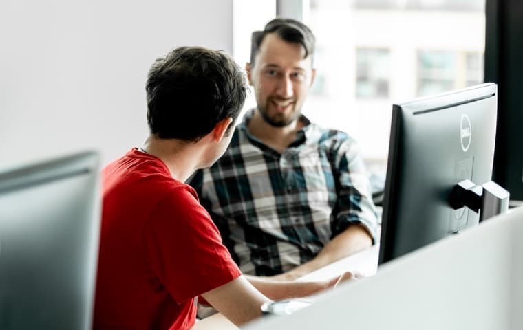 Two men engaging in a conversation at a work desk, with computers in the background. One wears a red shirt, the other a plaid shirt.