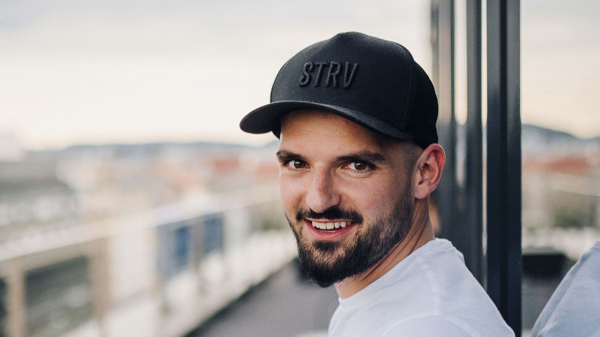 A man with a beard and a black cap smiles at the camera, with a blurred cityscape in the background.