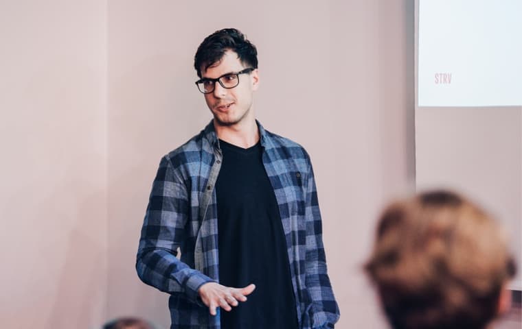 A man in glasses wears a plaid shirt, engaging an audience while presenting on a screen.