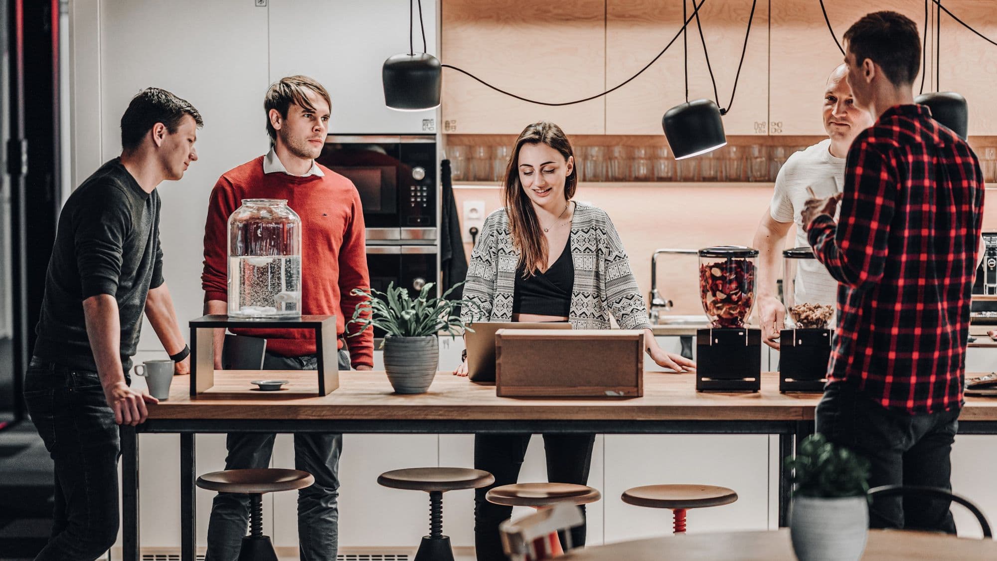 Five people engage in conversation around a kitchen island, with kitchen appliances and plants visible in a modern space.