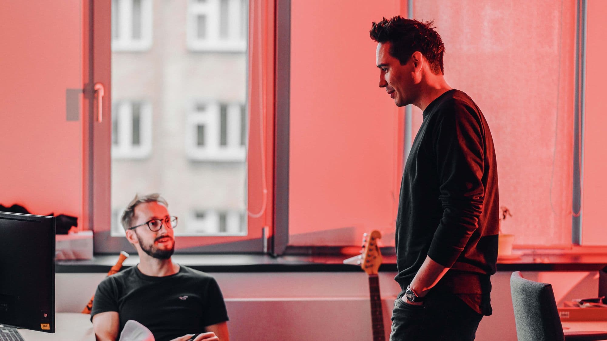 Two men converse in a brightly lit office space, with red-tinted windows and a guitar visible in the background.