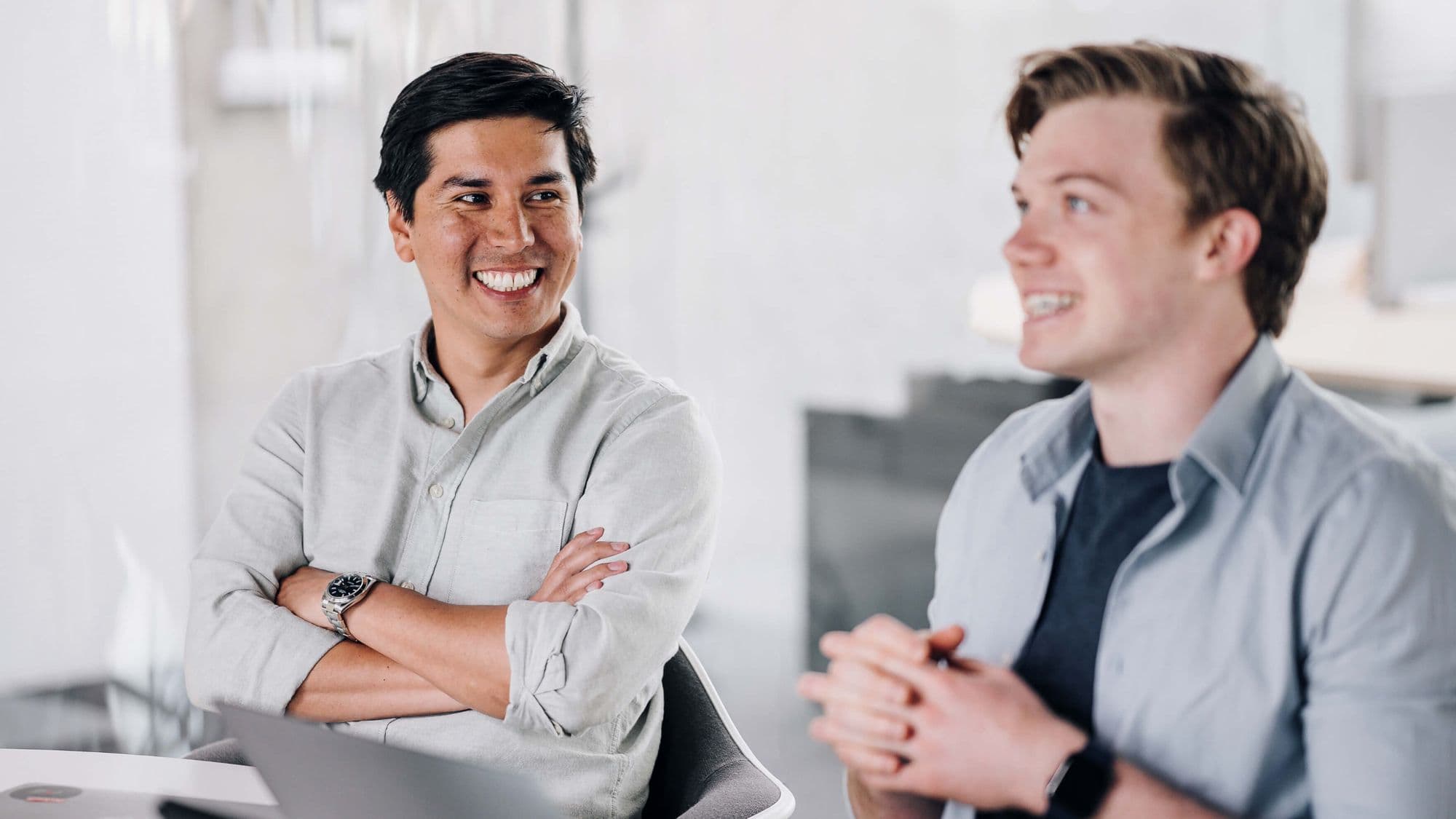 Two young men in a modern office setting engage in a friendly conversation, both smiling and appearing relaxed.