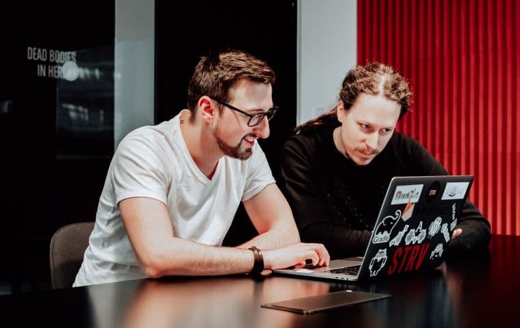 Two men collaborating on a laptop, surrounded by colorful wall panels and a modern workspace.
