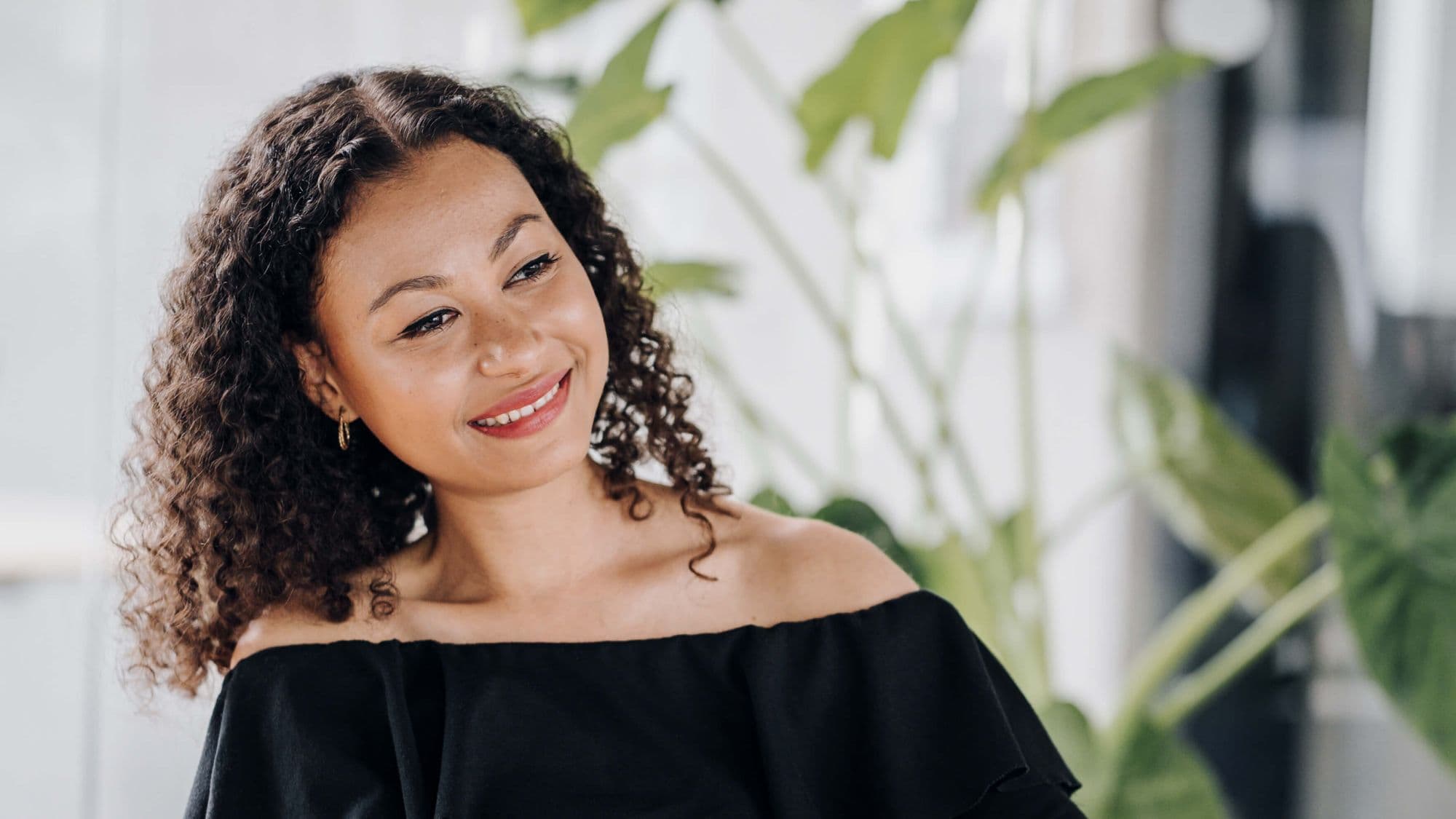 A woman with curly hair smiles while sitting in front of green plants, wearing a black off-the-shoulder top.
