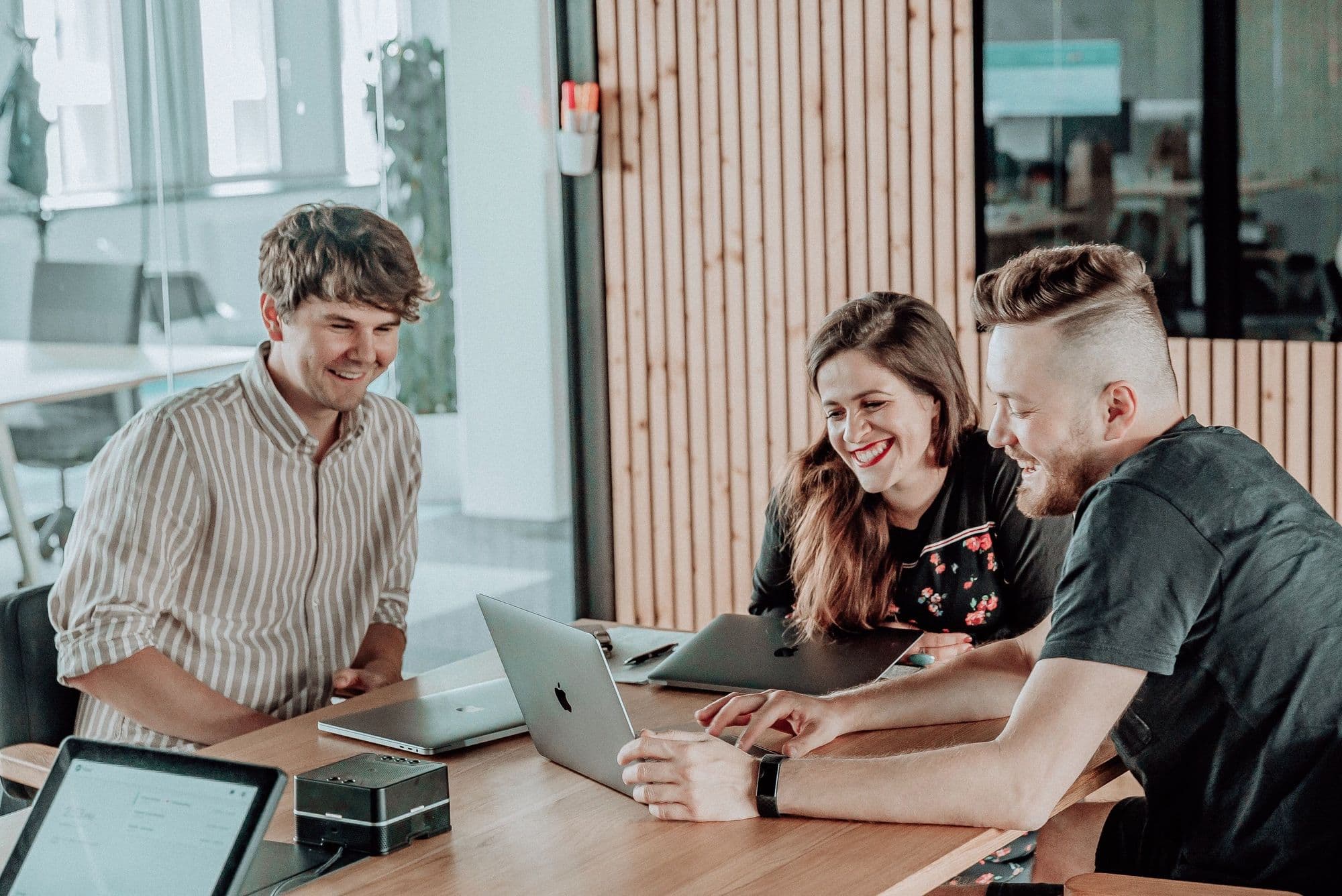 Three colleagues share a light moment while collaborating on a laptop in a modern office setting.