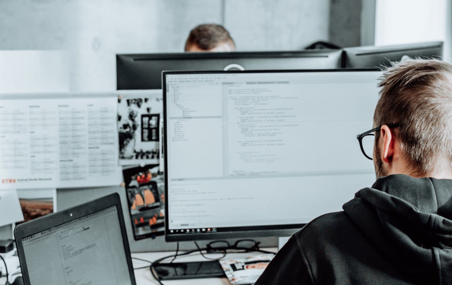 A person with glasses works at a desk with multiple computer monitors displaying code, papers, and notes in a modern office.