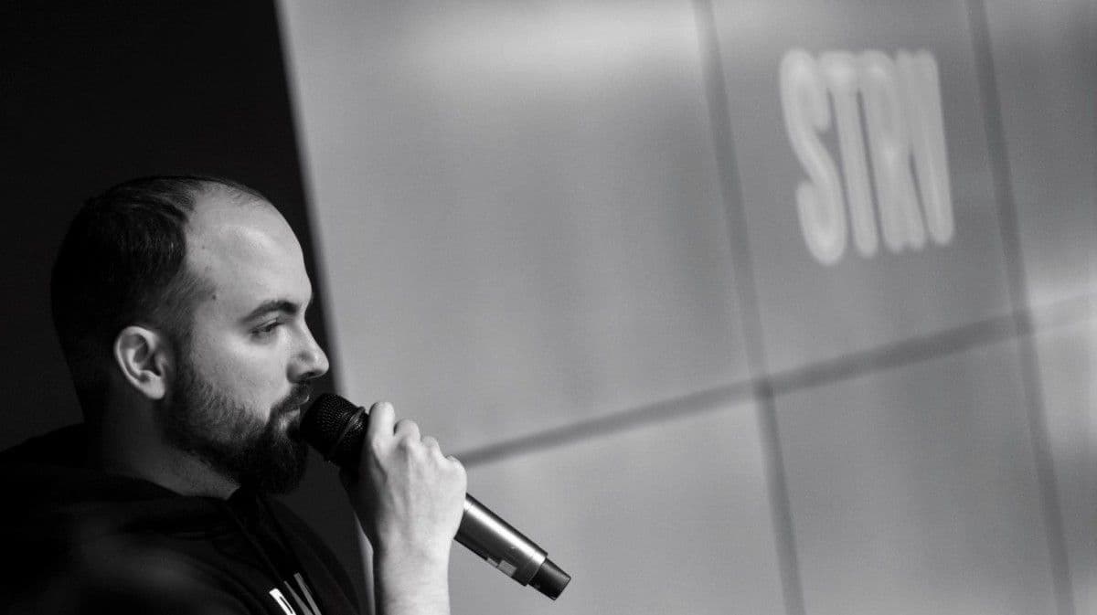A man with a beard speaks into a microphone, with "STN" visible in the blurred background.