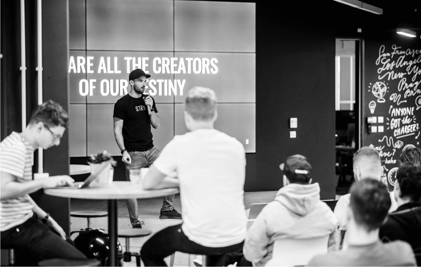 A speaker stands on stage addressing an audience, with a screen behind displaying the text, "ARE ALL THE CREATORS OF OUR DESTINY."