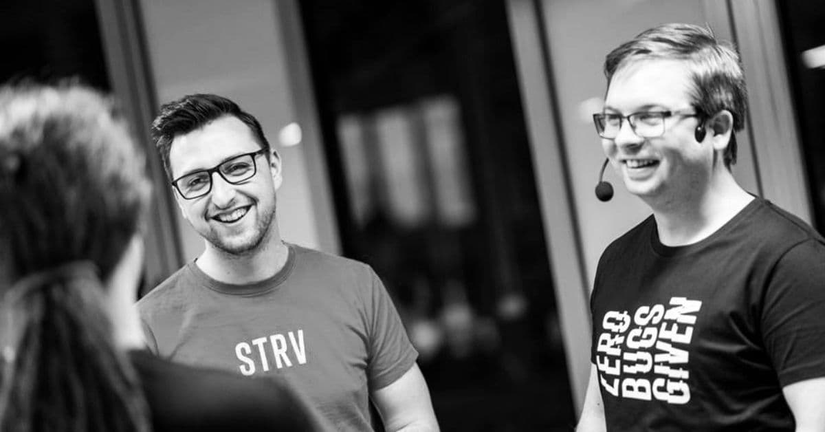 Two men in casual attire, smiling and engaging in conversation, one wearing a headset, in a modern office setting.