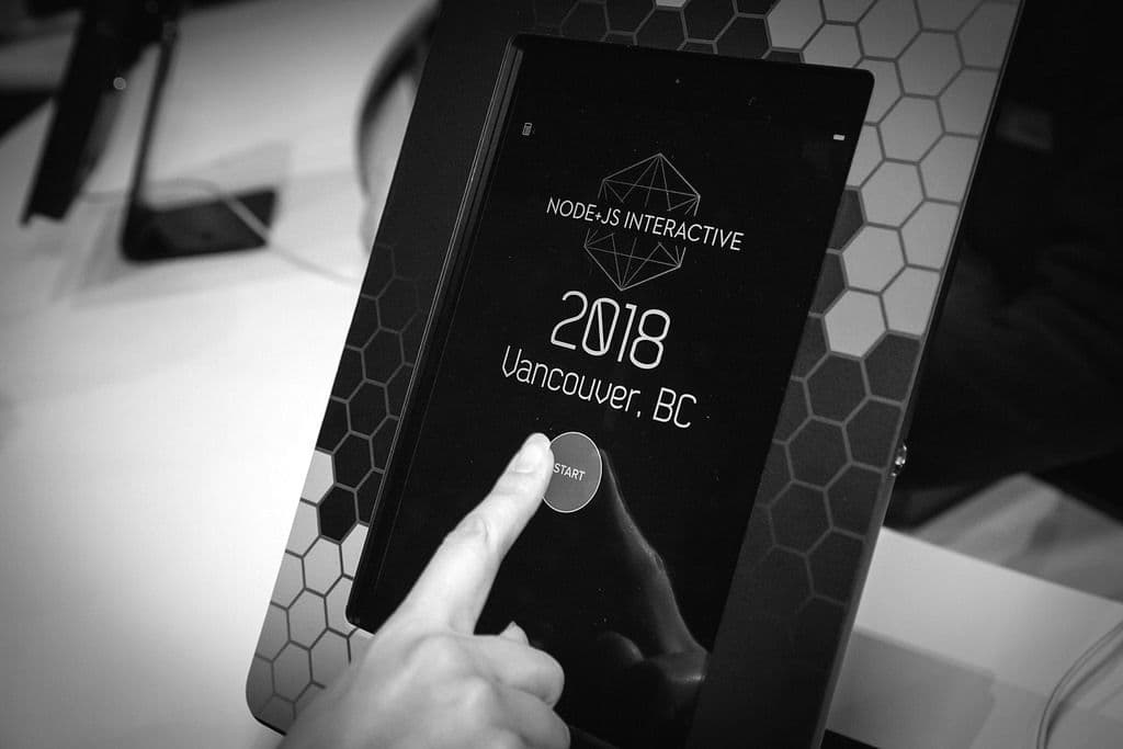 A hand touching a touchscreen displaying "Node.js Interactive 2018" and "Vancouver, BC" with a "START" button.