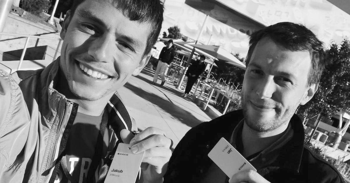Two men smiling at the camera, holding up name tags while standing outdoors with others in the background.