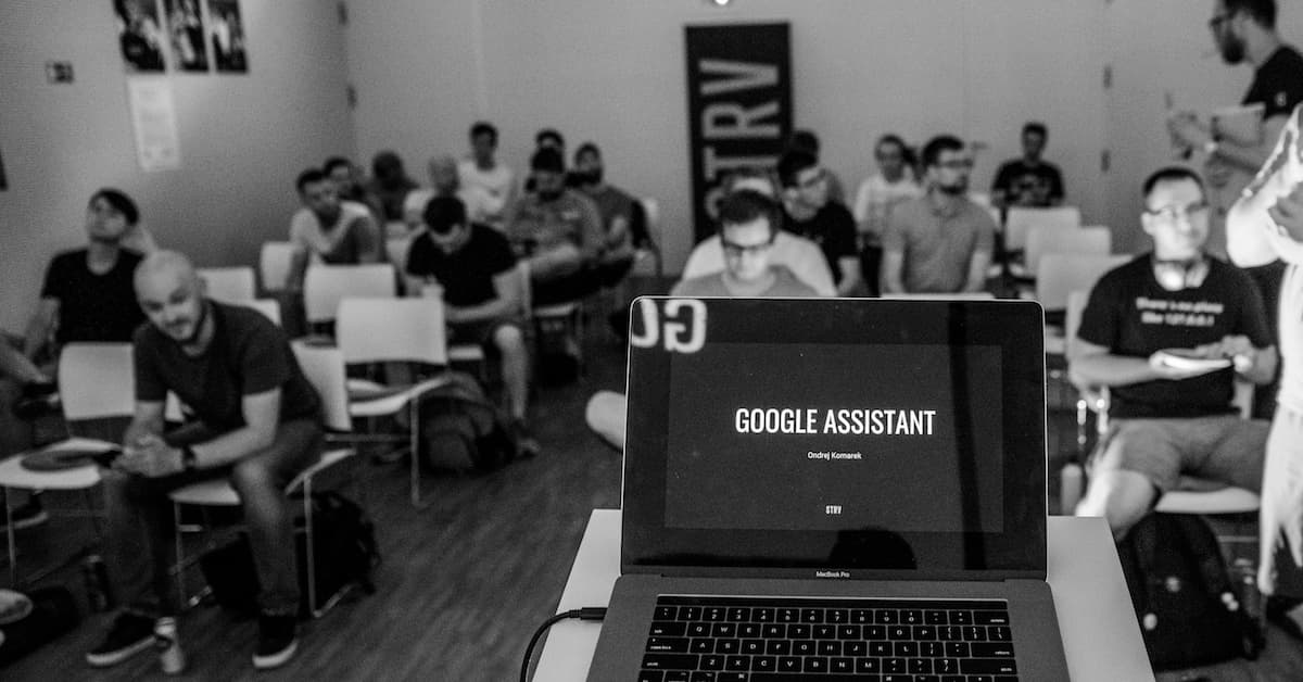 A laptop displaying "GOOGLE ASSISTANT" and the speaker's name, with an audience seated in a dim room.