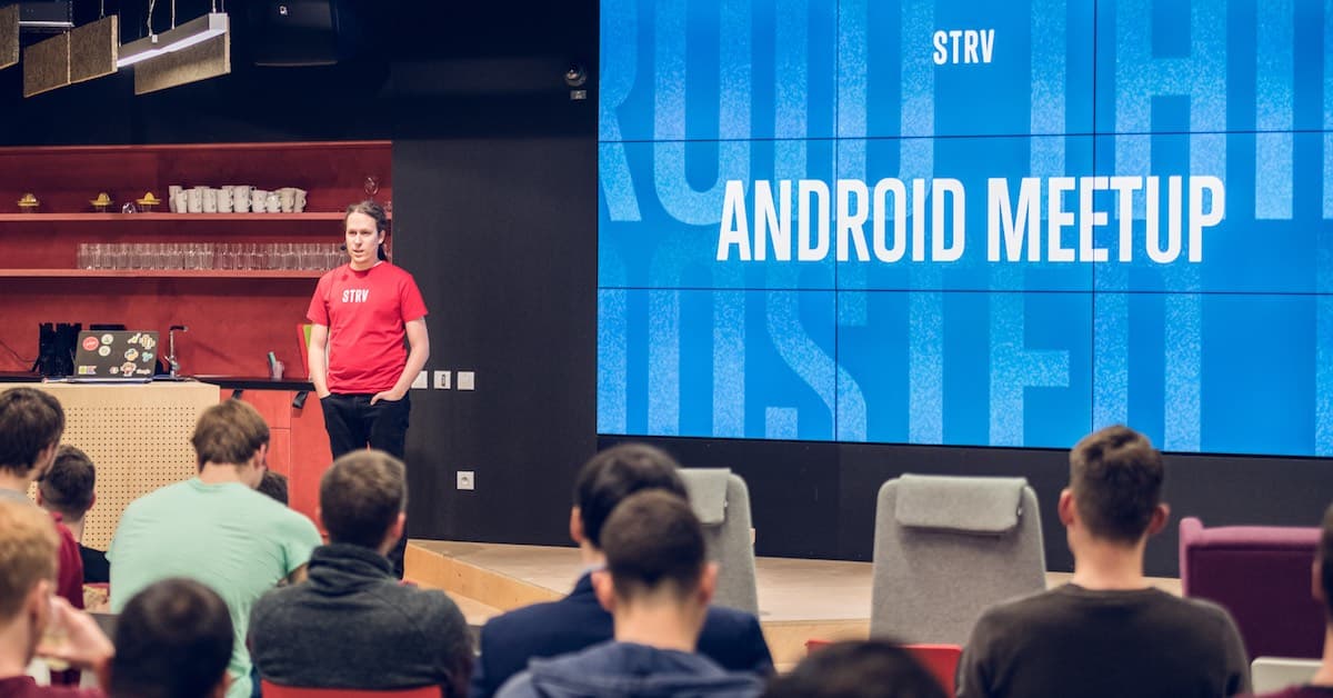 A speaker stands in front of an audience at an Android meetup, with a large screen displaying "ANDROID MEETUP."