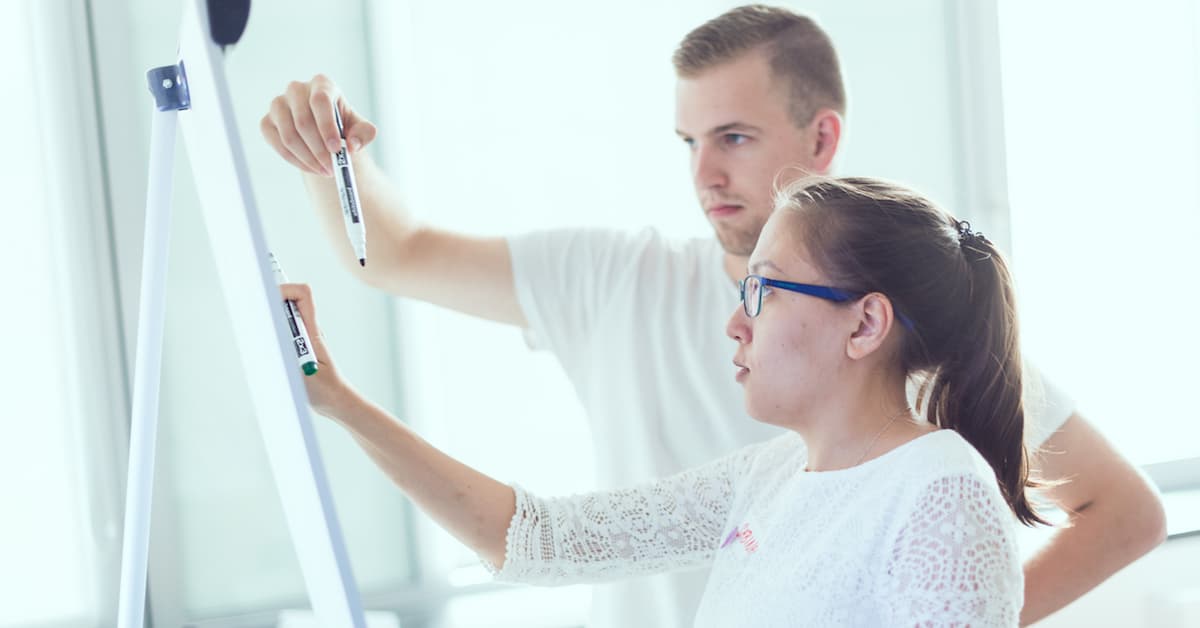 Two people, one writing on a whiteboard with a marker, while the other observes and discusses. Light-filled environment.