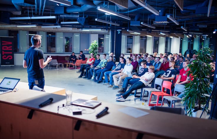 A speaker presents to an audience in a modern workspace, with a variety of chairs and plants in the background.