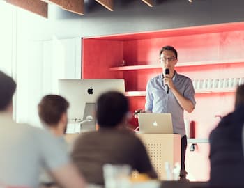 A speaker stands at a podium with a microphone, addressing an audience in a modern, well-lit room.