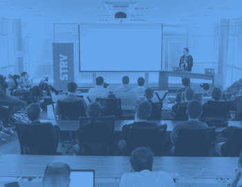 A presenter stands at a podium addressing an audience seated in a classroom with a large screen displaying content.