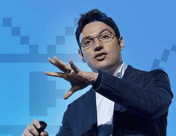 A man in a dark jacket gestures while speaking, with a blue background featuring pixelated graphics.