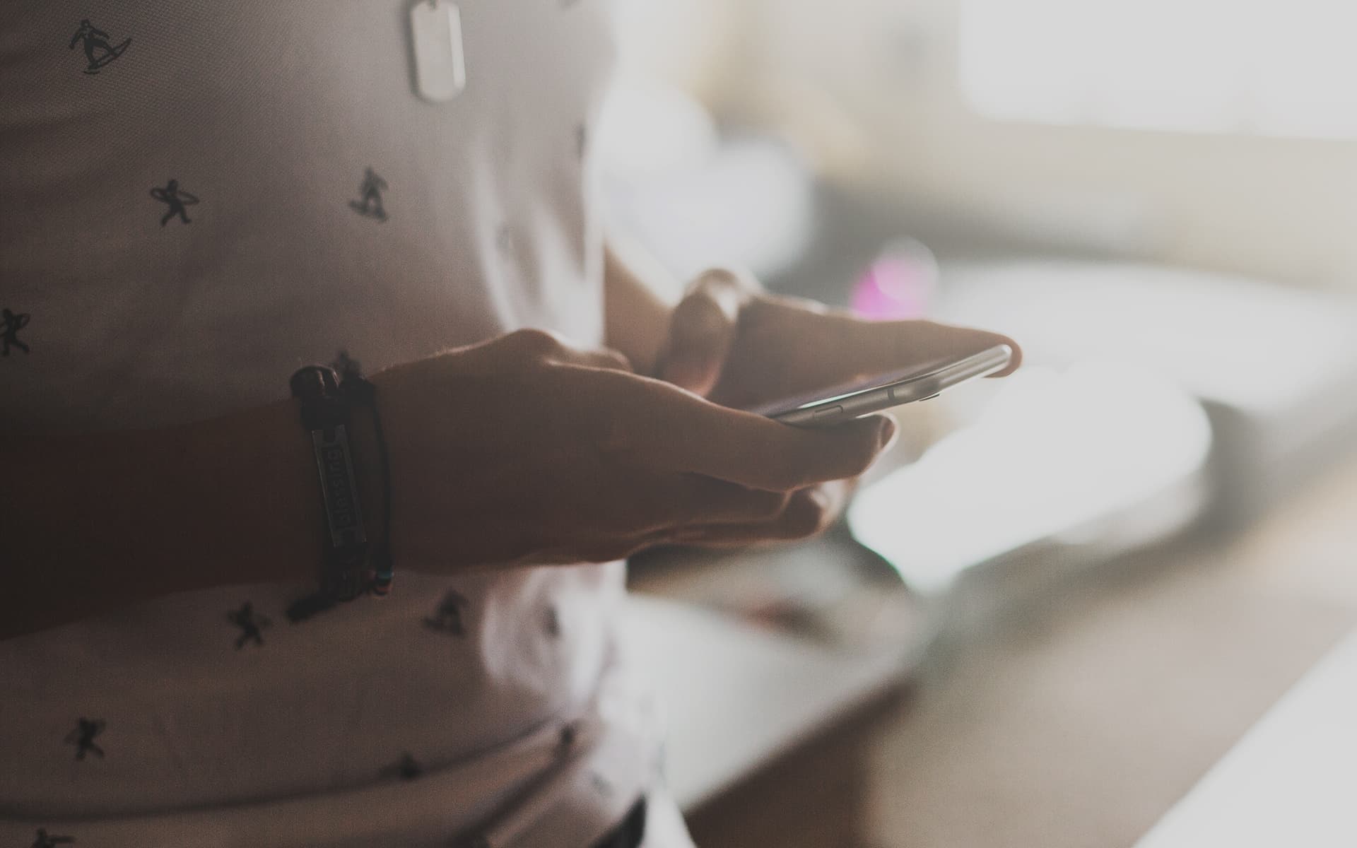 A person holding a smartphone, wearing a patterned shirt and a wristband, in a softly lit environment.