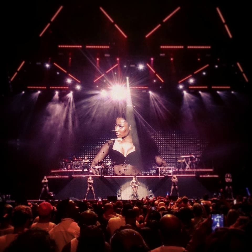 A performer in a black outfit stands on stage with bright lights, surrounded by dancers and an energetic audience.