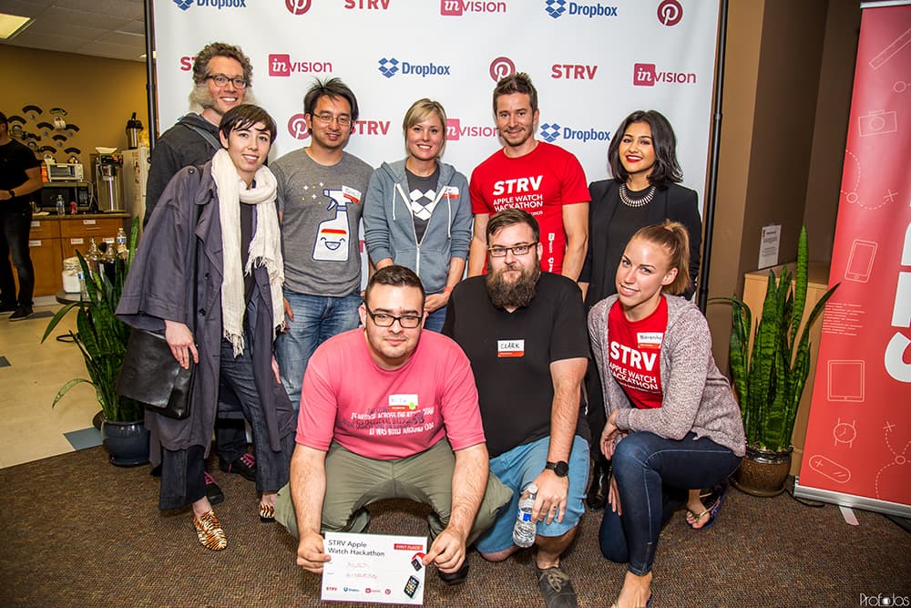 A group of eight people poses for a photo at an event, holding a sign that says "FIRST PLACE" in front of a branded backdrop.