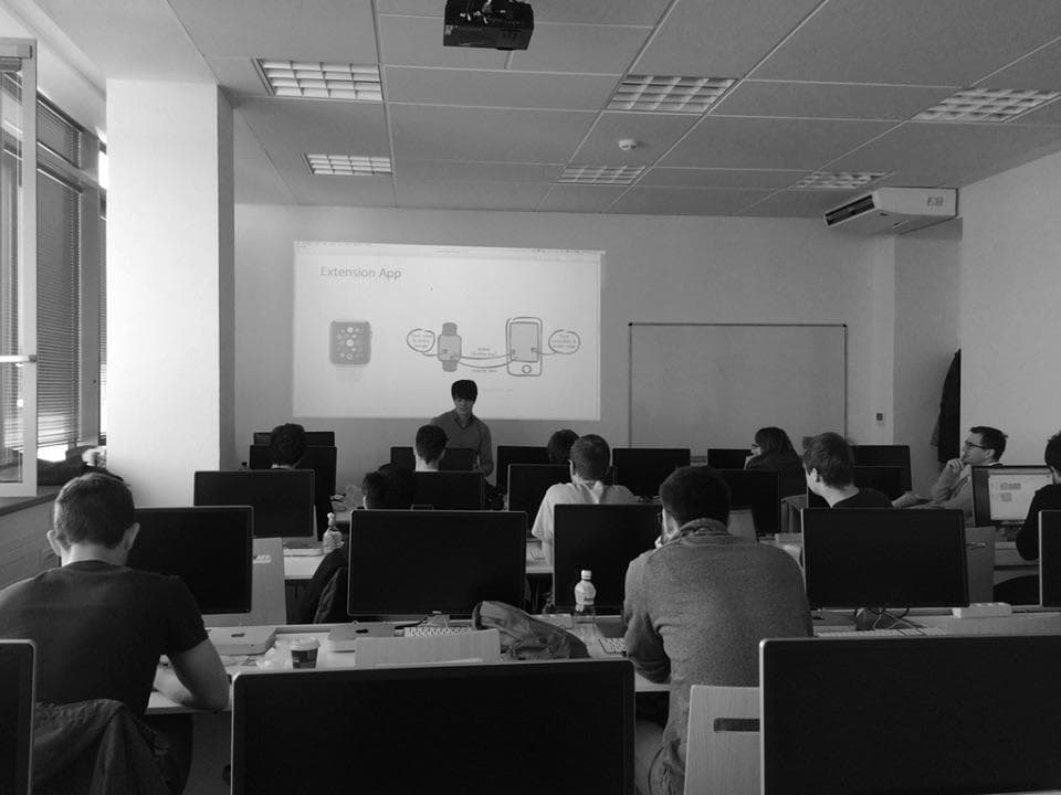A presenter discusses an "Extension App" in front of an audience seated at computers in a classroom.