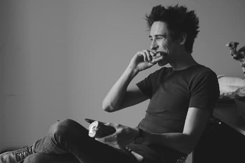 A young man with tousled hair is sitting and munching on something while holding a phone in his other hand.