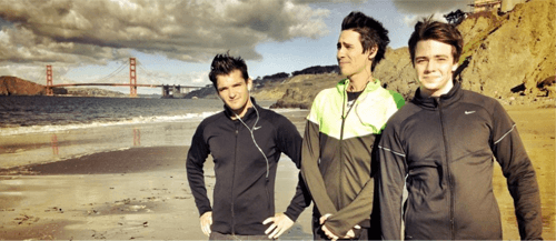 Three men in athletic jackets stand on a sandy beach with the Golden Gate Bridge in the background, under a cloudy sky.