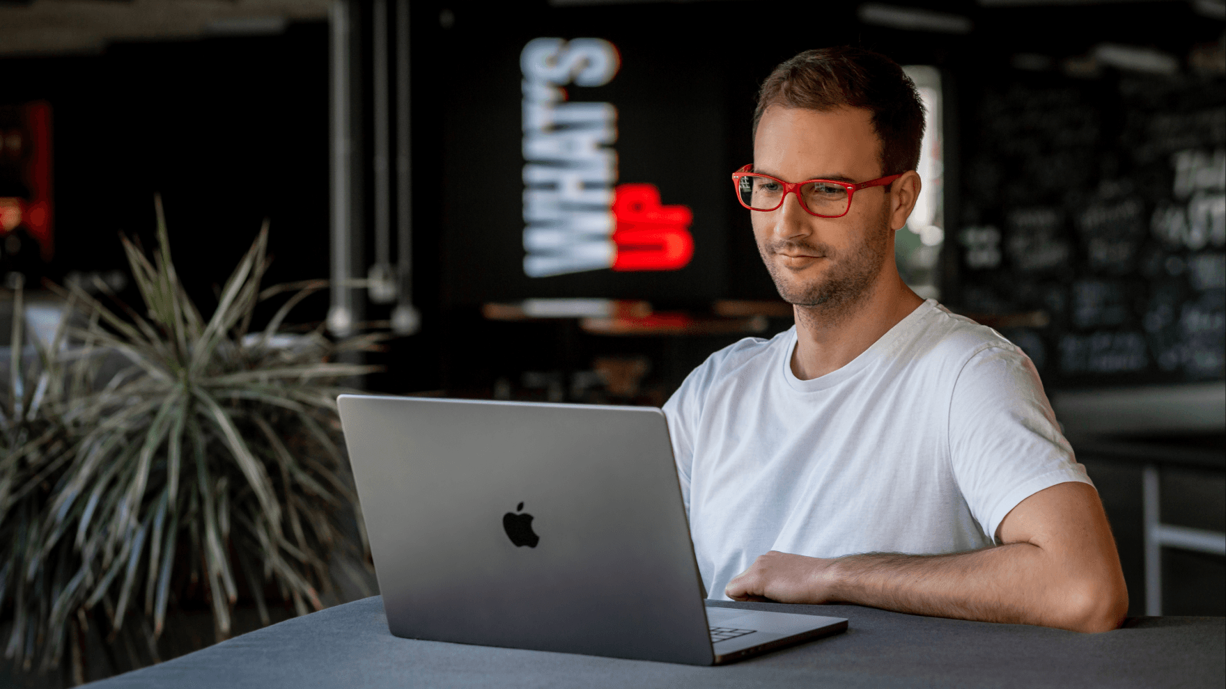 A man with red glasses sits at a table, focused on his laptop, with a "What's Up" sign illuminated in the background.