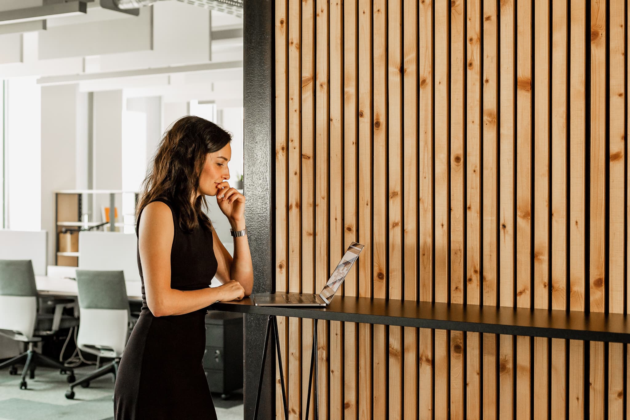 A woman in a black dress is standing at a table, focused on a laptop, with a wooden wall in the background.