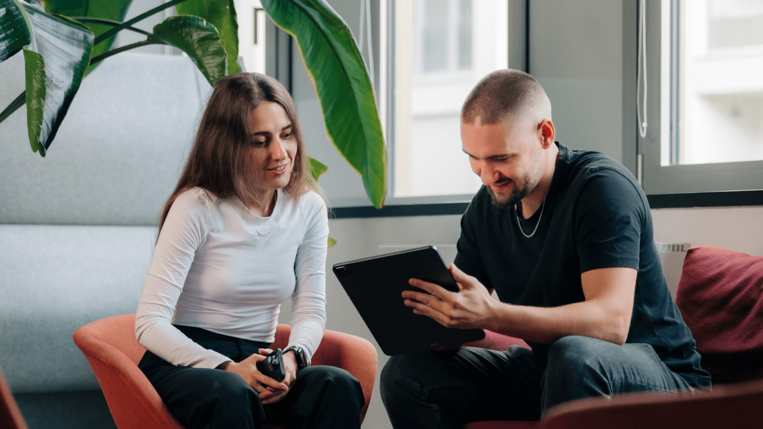 Two people sit in a cozy setting, engaging in conversation while looking at a tablet. A large plant adds to the atmosphere.